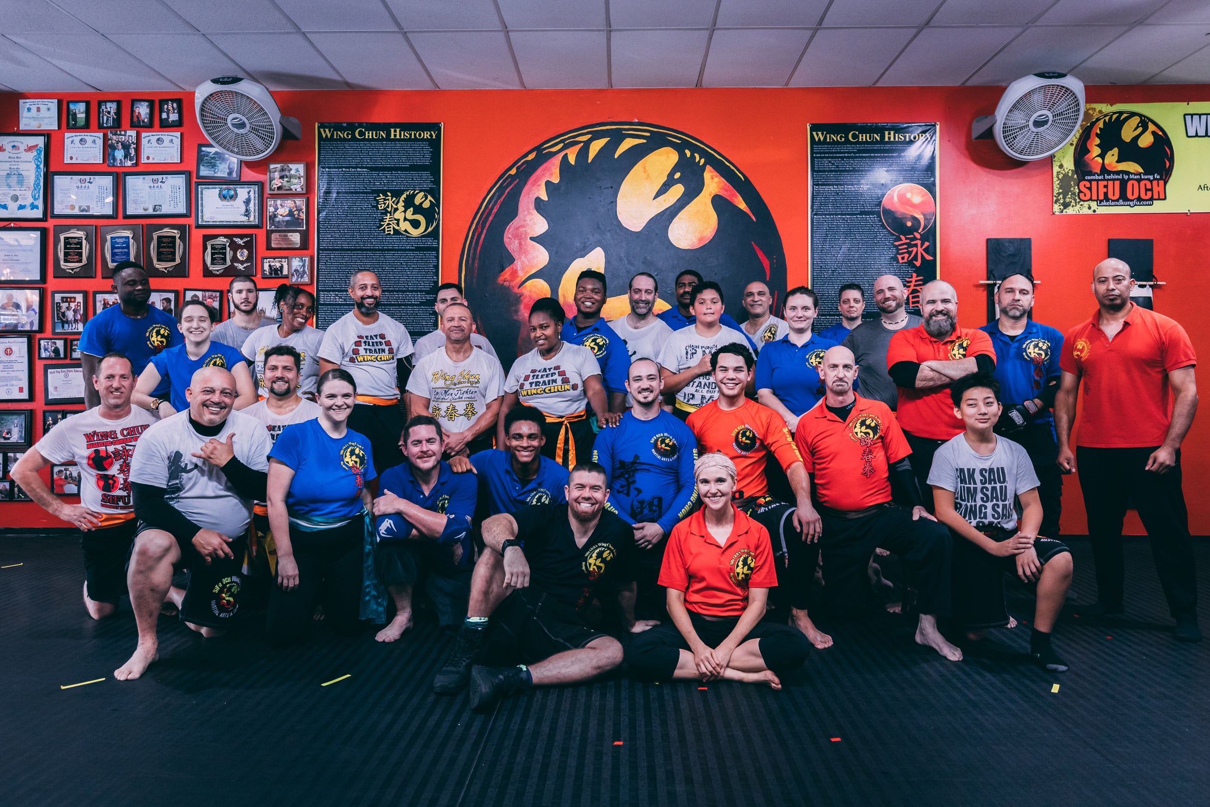 group photo of martial arts wing chun self defense practitioners sitting on workout mats in front of a large martial arts icon